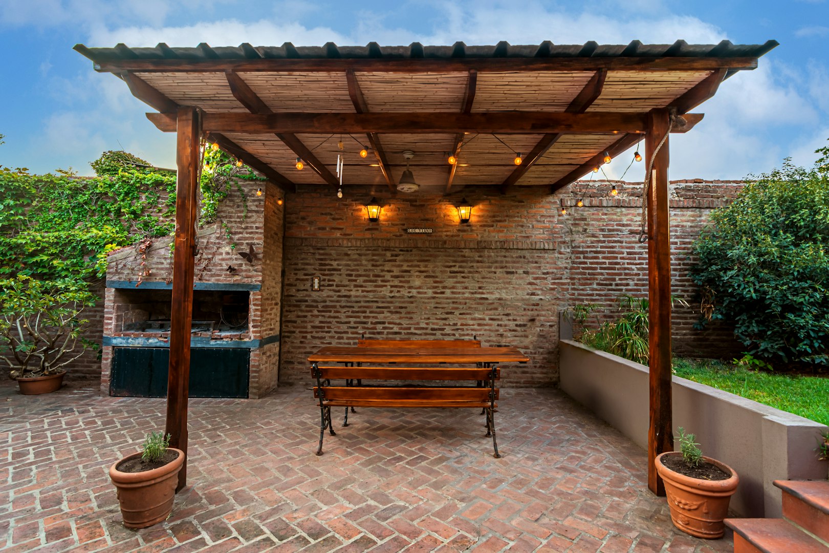 A covered outdoor patio with a brick barbecue, rustic wooden pergola, and brick flooring, surrounded by greenery — showcasing high-quality residential masonry work.
